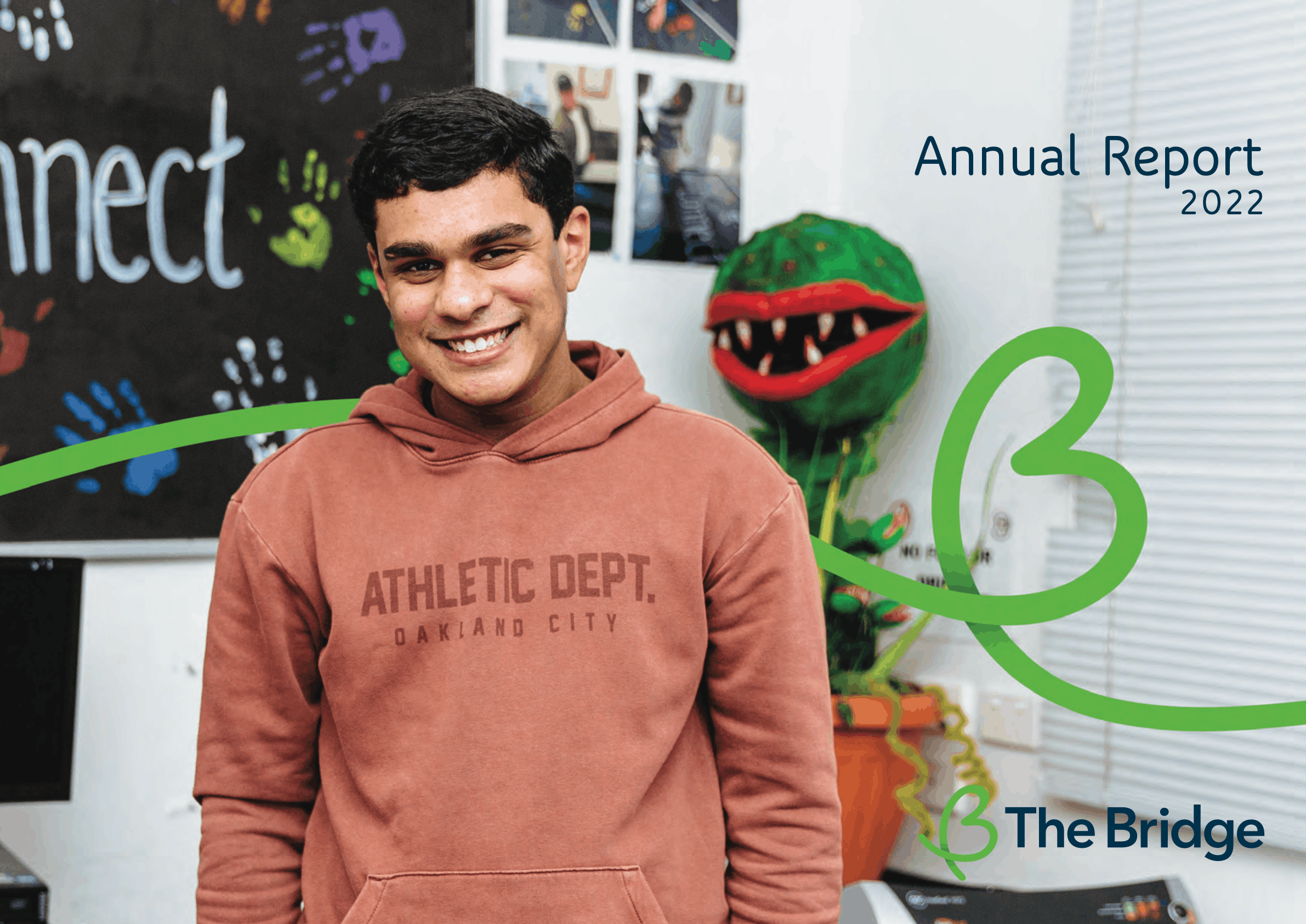 Young man smiling in orange hoodie, with a plant prop and handprint art behind him. The Bridge Annual Report 2022 cover.