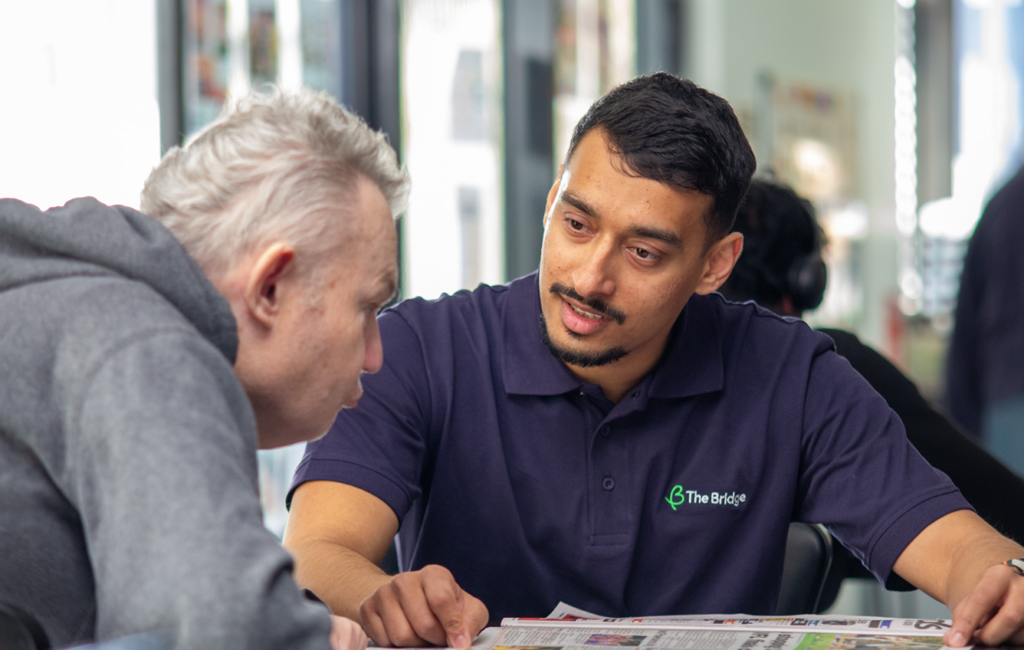 Man in a navy 'The Bridge' polo shirt talks with an older man in a grey hoodie over a document.