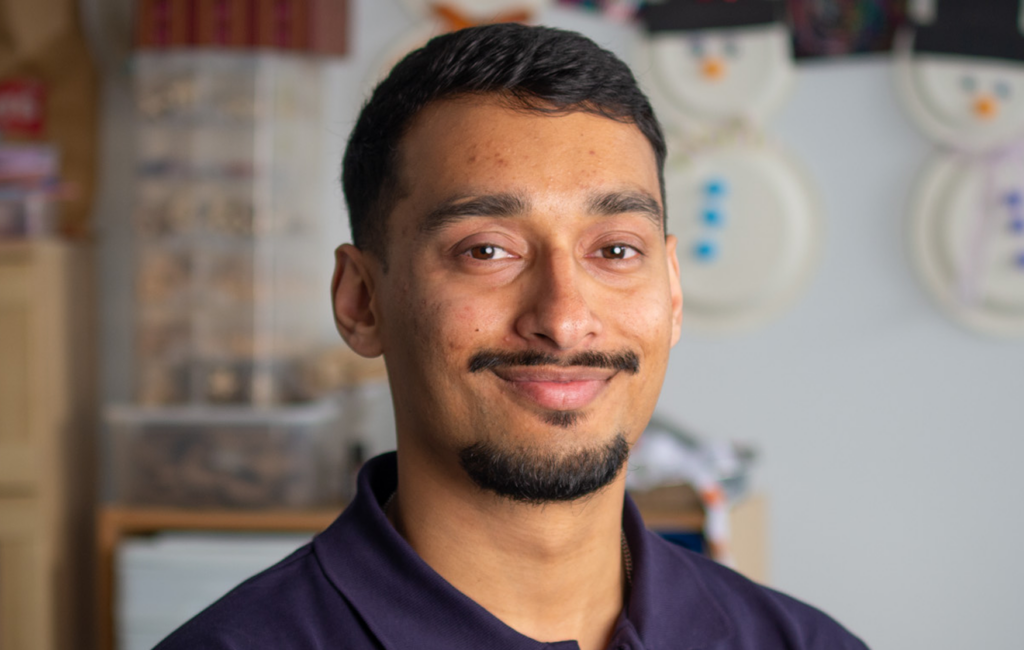 Man with short hair and moustache smiles in a navy polo shirt, snowman decorations on wall behind him.