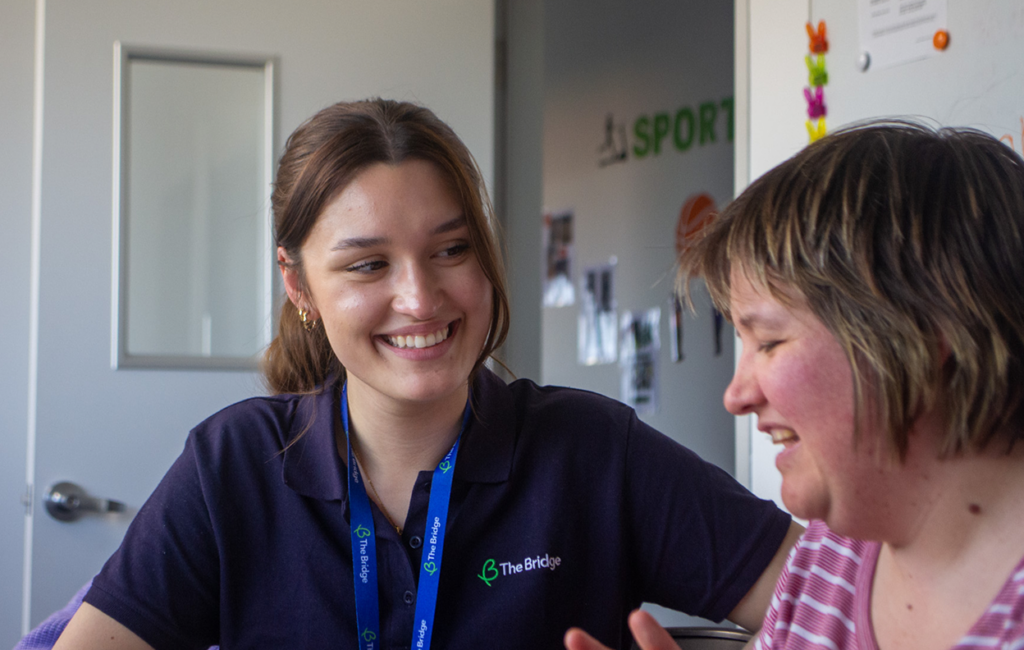 Two women sit together indoors, smiling and engaged in conversation. One wears a navy shirt with a The Bridge logo and a blue lanyard; the other wears a pink top. They appear to be in a supportive environment.