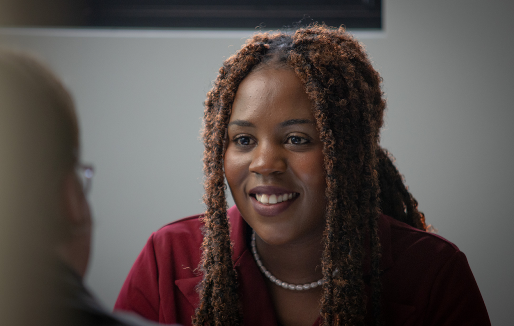 Woman with long braided hair, wearing a maroon top and pearl necklace, smiling in conversation with someone.