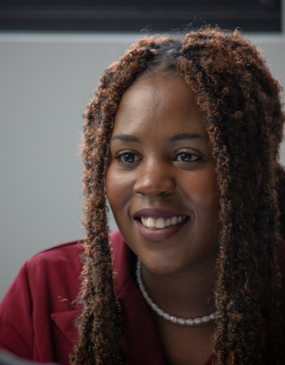 Woman with long braided hair, wearing a maroon top and pearl necklace, smiling in conversation with someone.