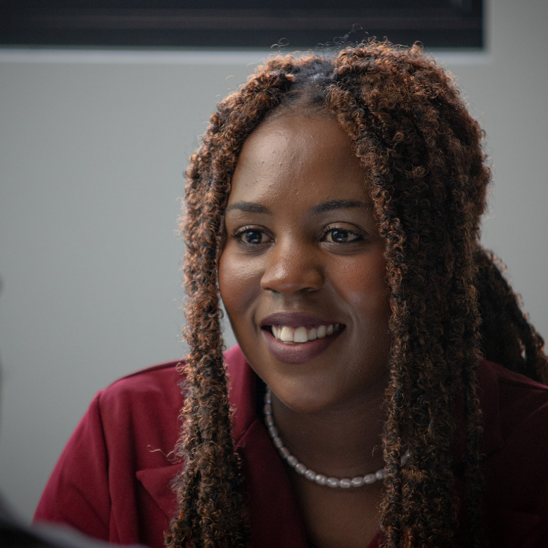 Woman with long braided hair, wearing a maroon top and pearl necklace, smiling in conversation with someone.
