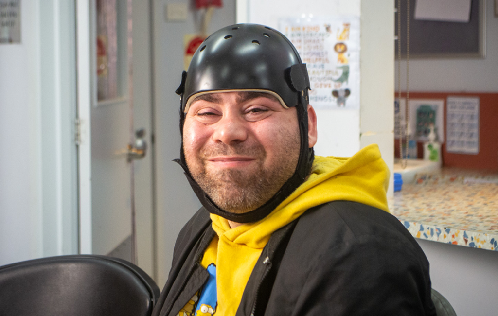 A man wearing a black protective helmet and yellow hoodie smiles at the camera while sitting indoors in a bright, casual setting.