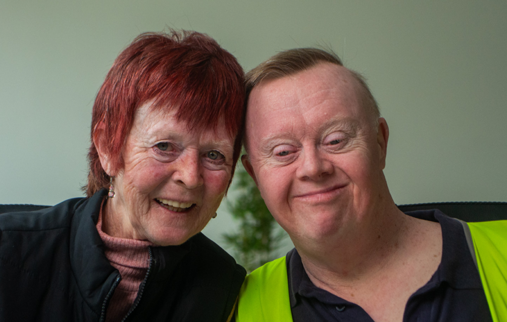 A woman with red hair and a man wearing a hi-vis vest smile closely together, heads touching.
