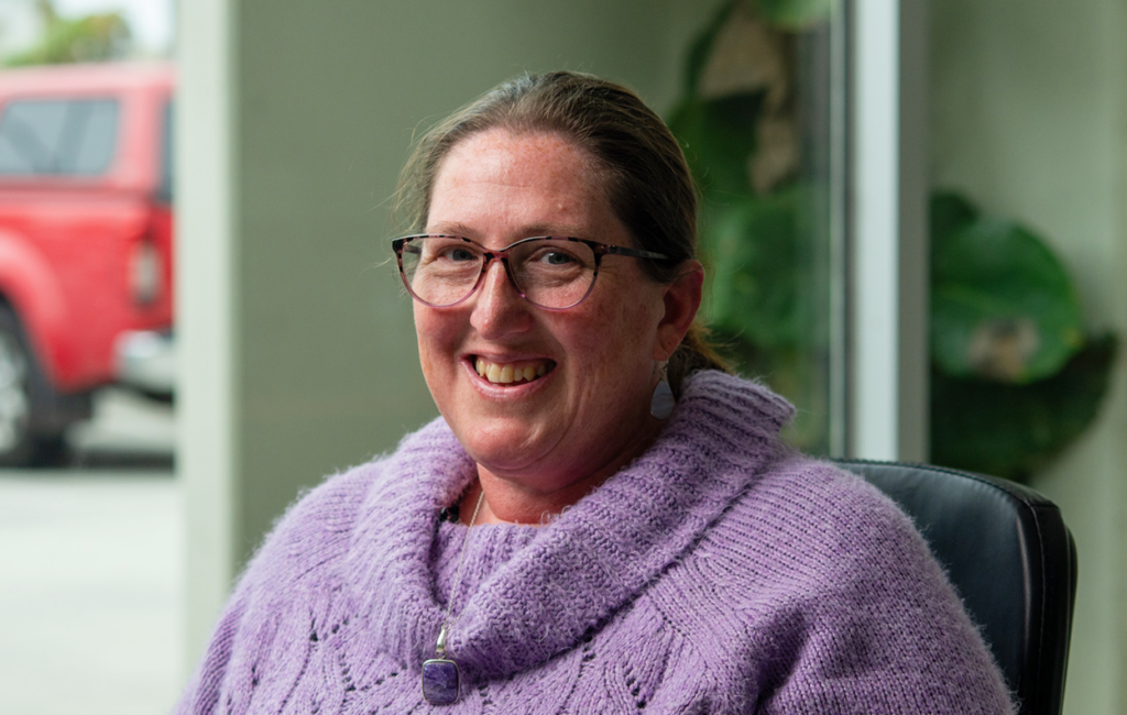 Woman with glasses wearing a purple knit jumper smiles, seated indoors near a doorway.