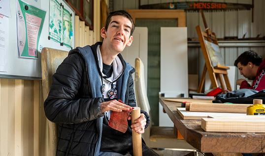 Young man sanding a wooden piece by hand, seated in a men's shed workshop with another person working behind him.