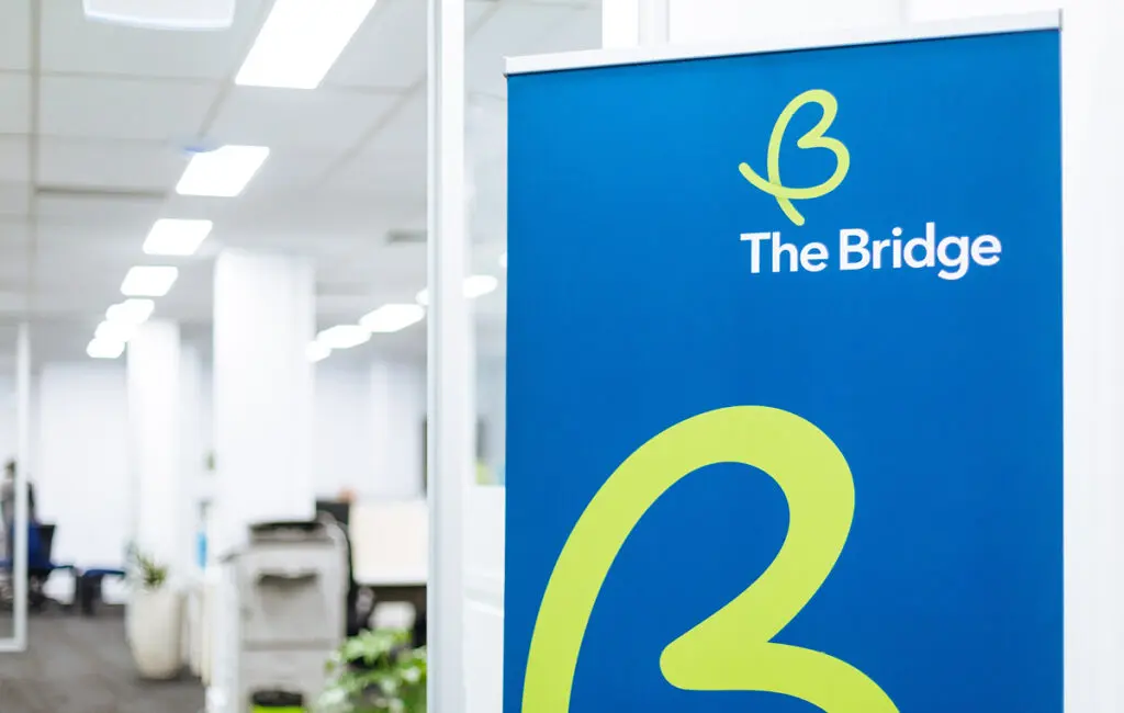 Blue pull-up banner with The Bridge logo, standing in an office with desks and plants.