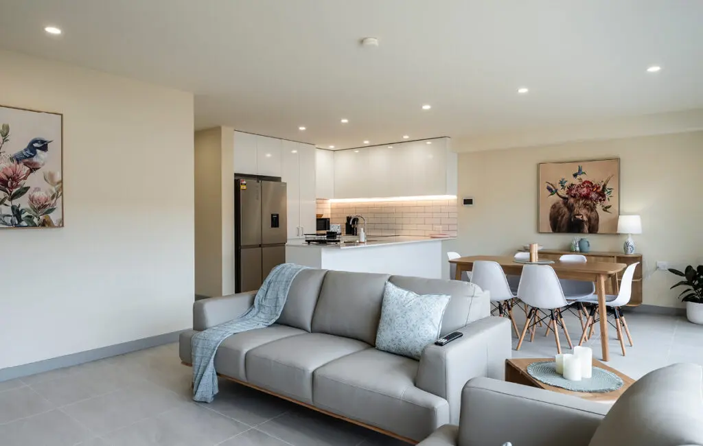 Open-plan living area with grey sofas, kitchen with white cabinetry, and dining table with white chairs.