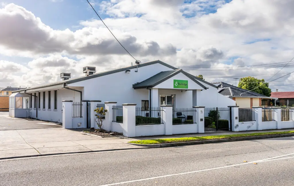 Single-storey white building with 'The Bridge' green sign, front fence and gate, on a street corner.