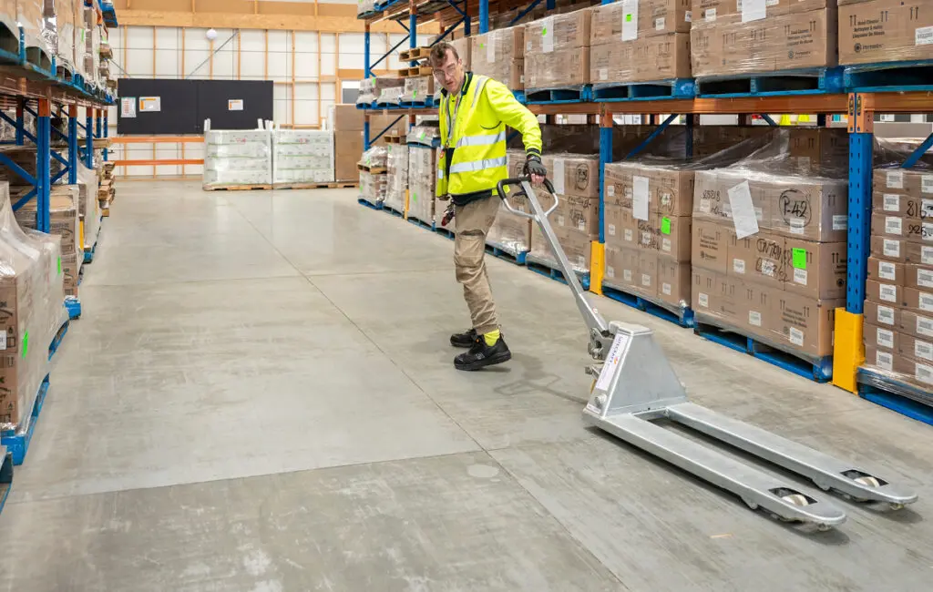 Man in high-vis vest pushes a manual pallet jack along an aisle in a warehouse with stacked pallets.