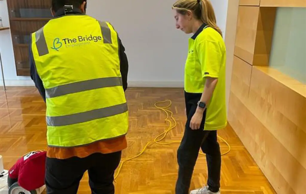 Two workers in yellow hi-vis, one wearing a Bridge Employment vest, stand by a floor buffer on wood flooring.