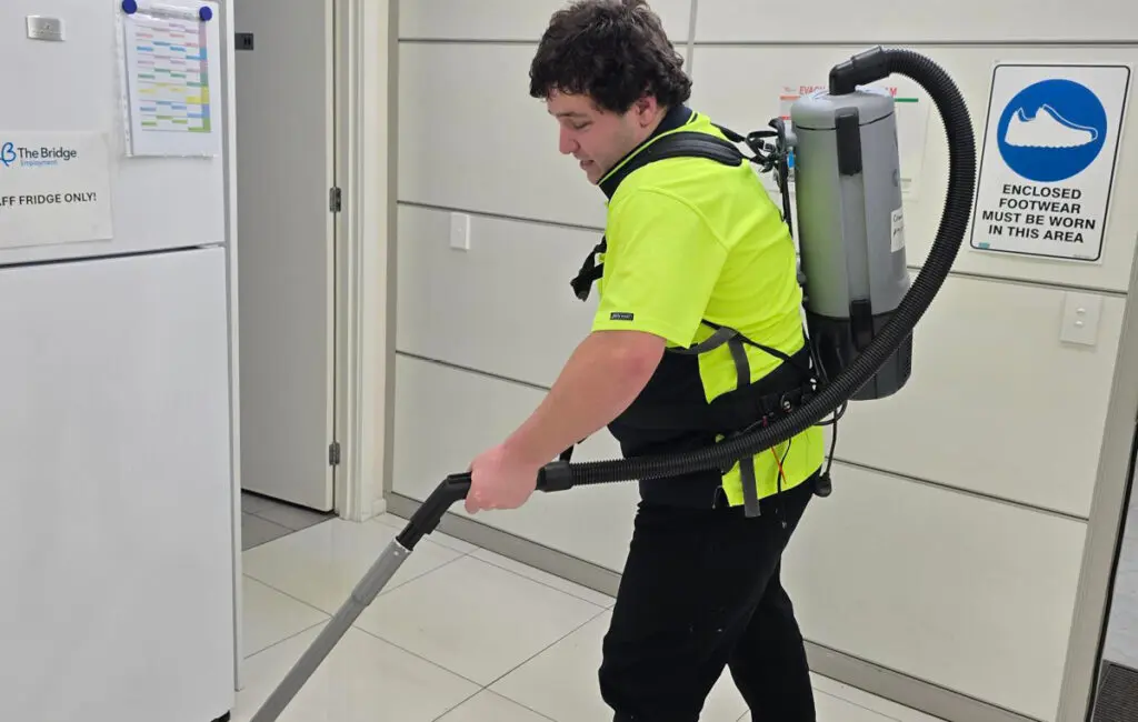 Man in high-vis shirt vacuums tiled floor near fridge, wearing a backpack vacuum cleaner.
