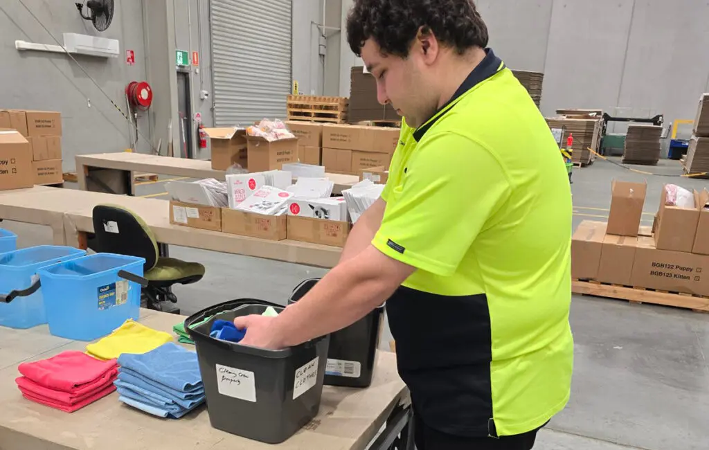 Man in a hi-vis shirt sorts folded cloths into a labelled bin on a warehouse workbench.