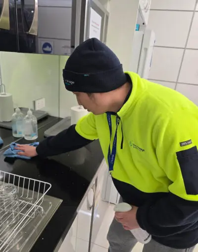 Man in hi-vis jacket and beanie wipes a kitchen benchtop with a cloth and spray bottle.