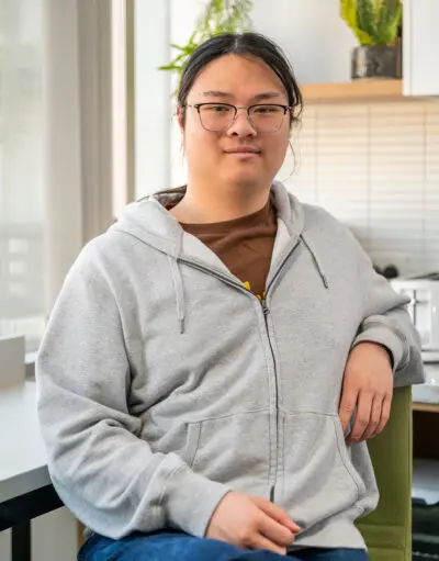 Person with glasses and grey zip-up hoodie sits smiling in a kitchen, plants and blinds in background.