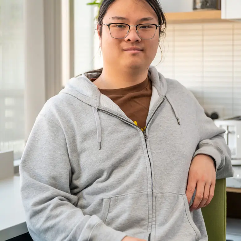 Person with glasses and grey zip-up hoodie sits smiling in a kitchen, plants and blinds in background.