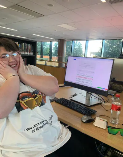 Person smiling with hands on cheeks at a desk, computer screen showing a completed quiz result behind them.