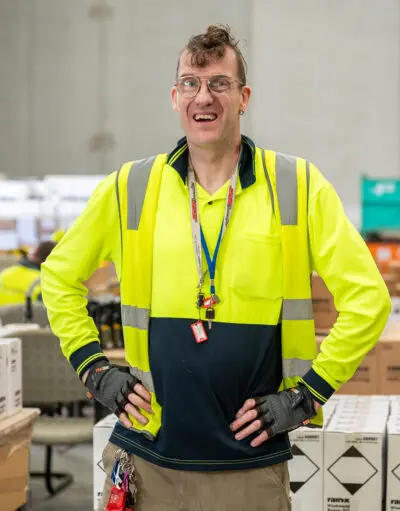 Man in hi-vis jacket, gloves and lanyard smiling with hands on hips in a warehouse with stacked boxes.
