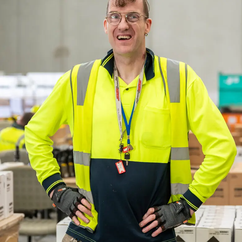 Man in hi-vis jacket, gloves and lanyard smiling with hands on hips in a warehouse with stacked boxes.