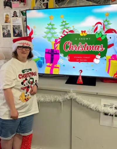 Person in Santa hat and Christmas T-shirt beside TV screen showing 'A Snowy Christmas' title slide