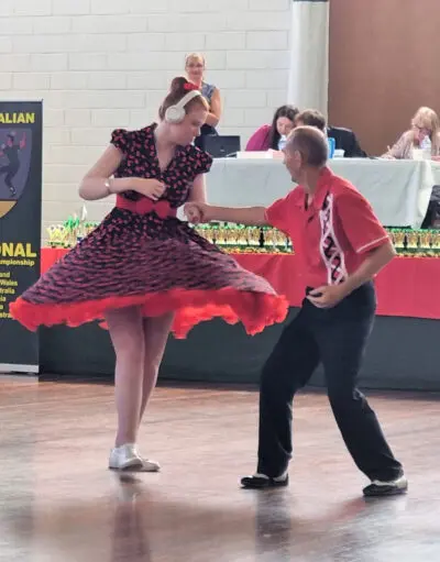 Woman wearing headphones in a red patterned dress dances with a man in a red shirt, judges seated behind them.