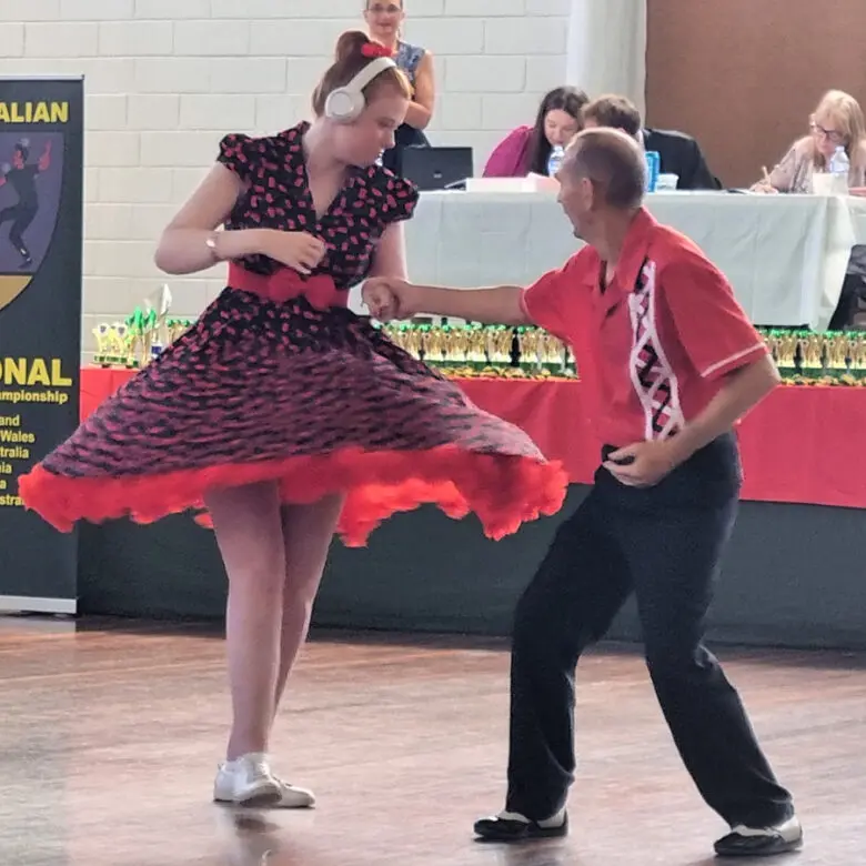 Woman wearing headphones in a red patterned dress dances with a man in a red shirt, judges seated behind them.