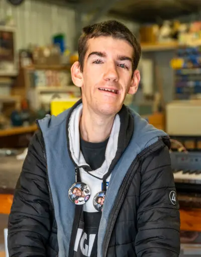 Man with badges on hoodie smiling at camera, standing in a workshop or shed.