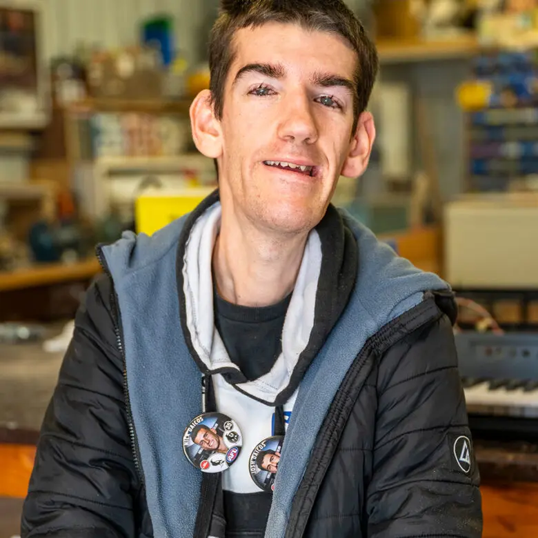 Man with badges on hoodie smiling at camera, standing in a workshop or shed.