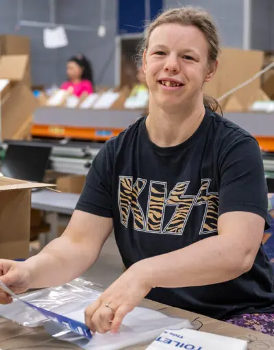 Woman in a KISS t-shirt smiles at camera while packing plastic bags at a warehouse workstation.
