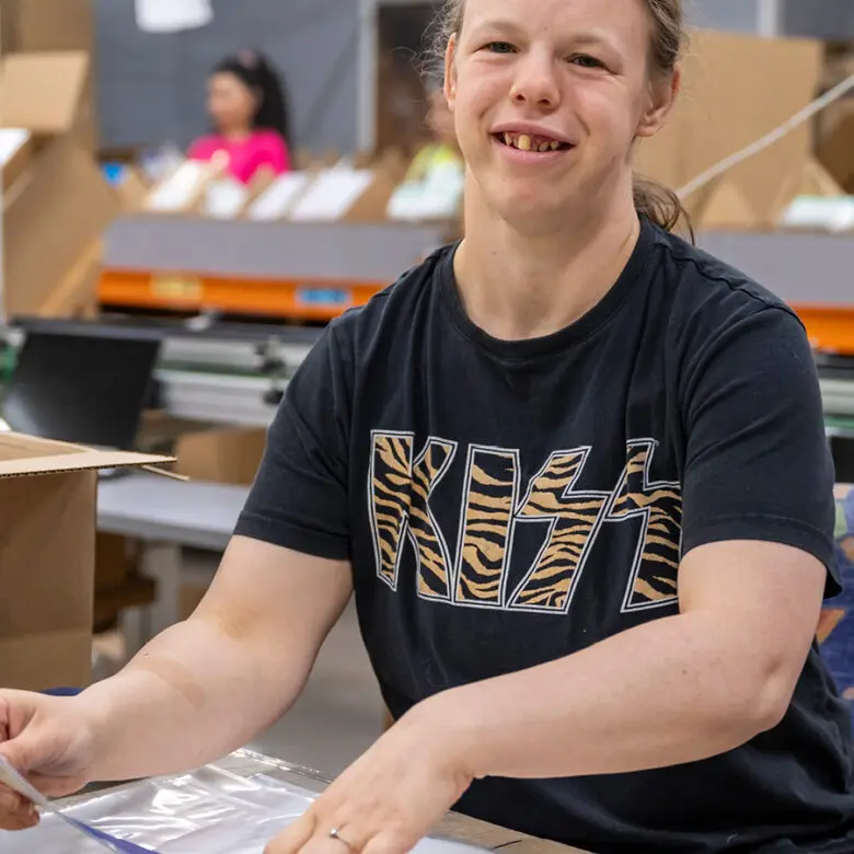 Woman in a KISS t-shirt smiles at camera while packing plastic bags at a warehouse workstation.