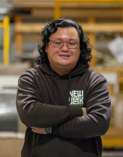 Man with glasses and shoulder-length hair, arms crossed, wearing a brown hoodie in a warehouse