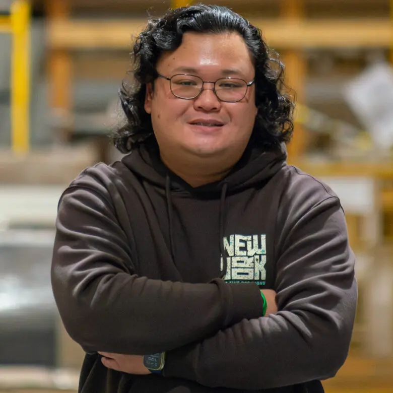 Man with glasses and shoulder-length hair, arms crossed, wearing a brown hoodie in a warehouse