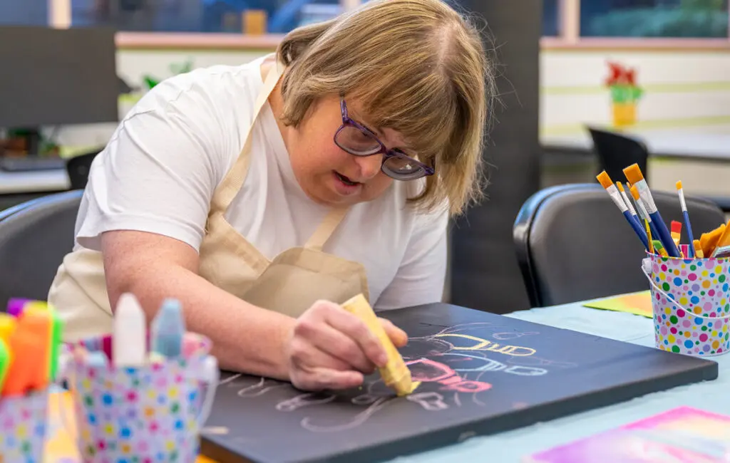 Woman wearing an apron and glasses draws with chalk on a black board at a table with paintbrushes.