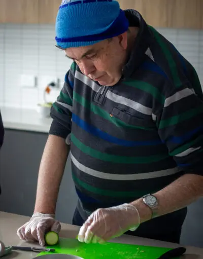 Man in a blue beanie and striped jumper wears gloves, slicing cucumber on a green board in a kitchen.