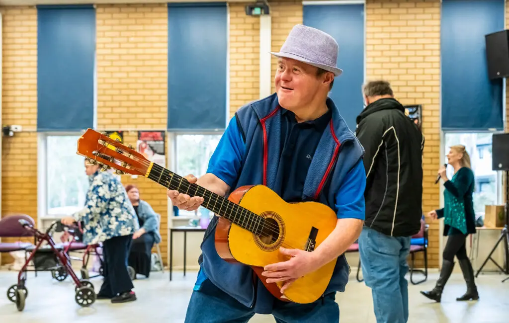 Man in a hat and blue vest plays acoustic guitar, smiling, with others in a community hall behind him.