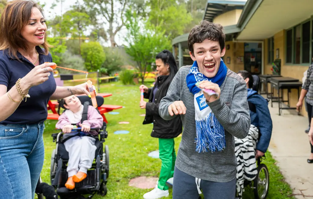 A young man wearing a scarf smiles broadly outdoors, with a woman blowing bubbles nearby and others in wheelchairs in a grassy courtyard.