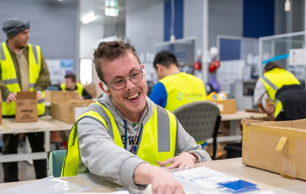 Man in glasses and hi-vis vest smiling while packing items at a table, with colleagues working in background.
