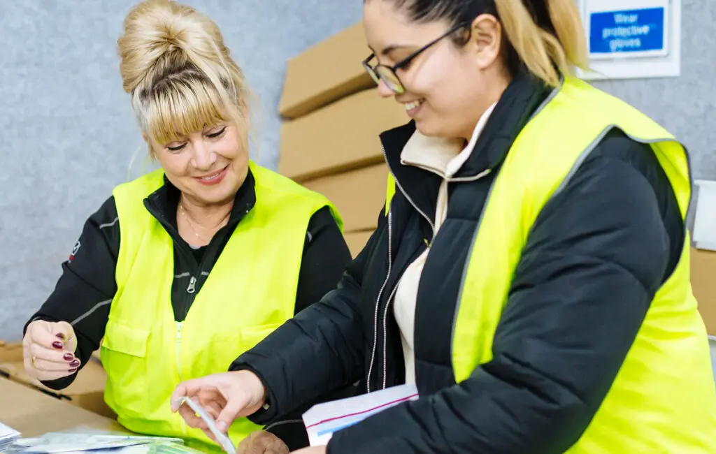 Two women in hi-vis vests smiling, checking documents at a table with cardboard boxes nearby.