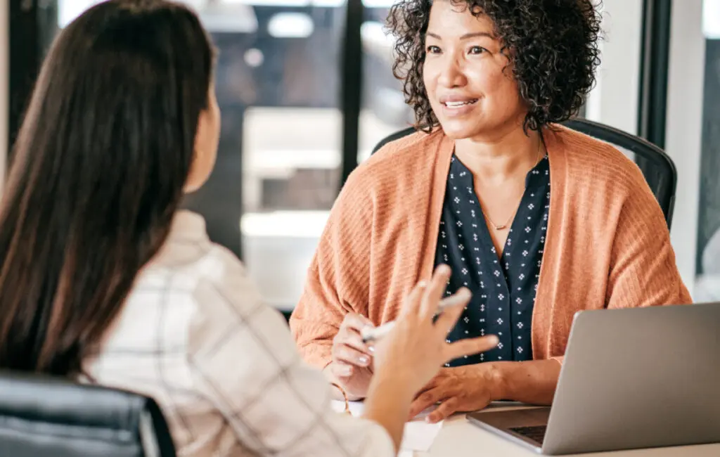 Two women in conversation at a table with a laptop, one smiling while listening to the other.