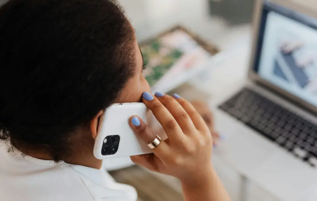 Woman holds a phone to her ear at a desk, with a laptop screen visible nearby.