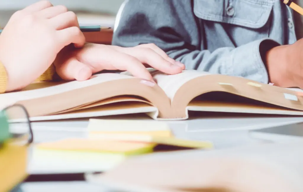 Two people's hands rest on an open book at a desk with sticky notes and pencils nearby.