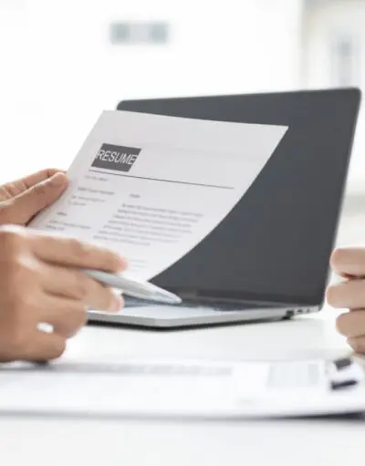 Person holding a resume and pen at a table, another person's clasped hands opposite, laptop between them