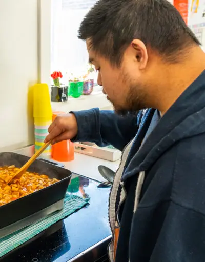 Man in a blue hoodie stirring a tomato-based dish with a wooden spoon in an electric frying pan.