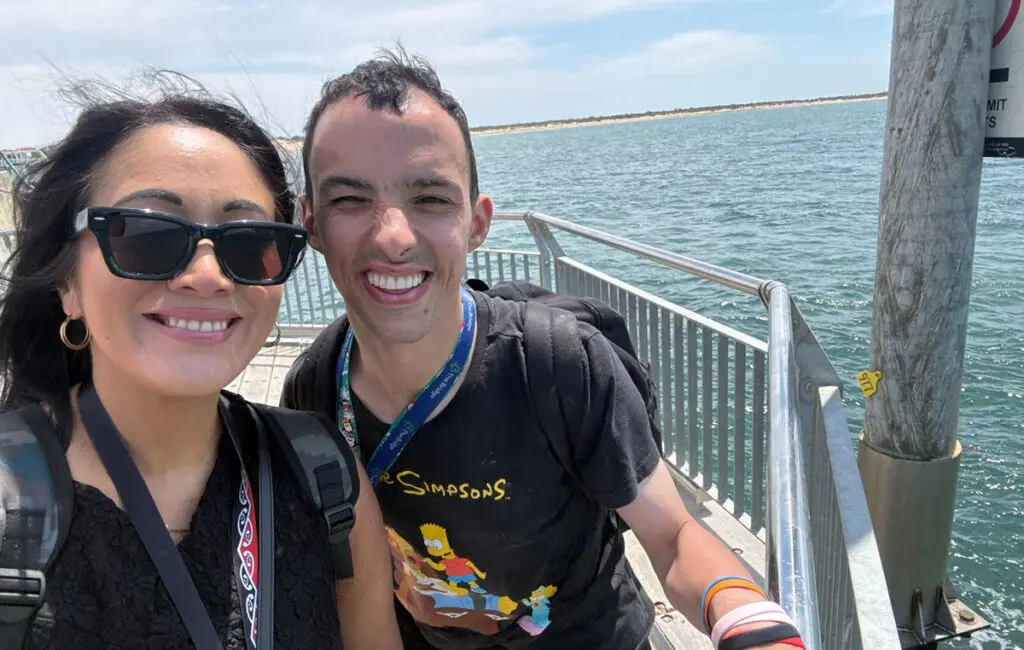 Woman with sunglasses and man wearing a Simpsons T-shirt smile on a pier beside the ocean.