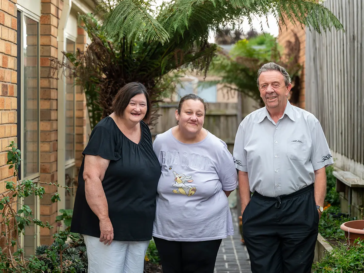Three people smiling side by side in a garden path beside a brick house and fern.