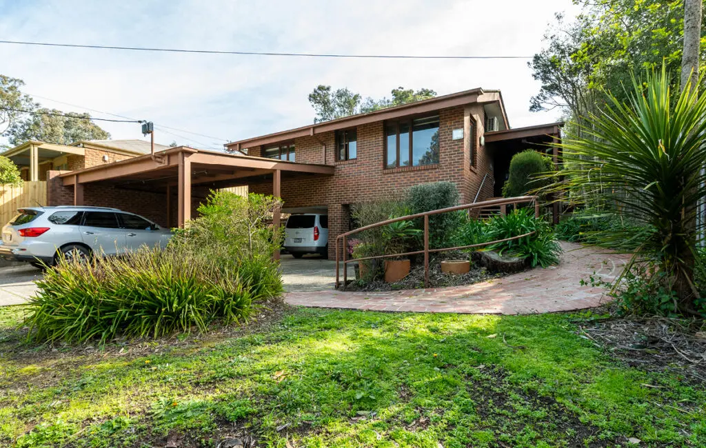 Brick house with carport, two cars parked, curved brick path with handrail, front garden and lawn