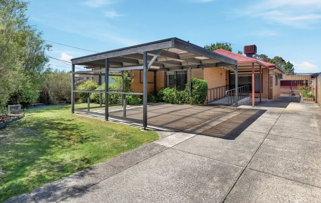 Brick house with red roof, carport, ramp with handrail to front entrance, and concrete driveway.