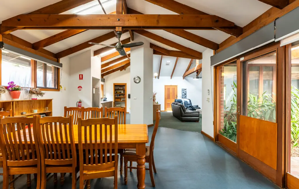 Open-plan room with wooden dining table and chairs, exposed beams, and lounge area visible beyond.
