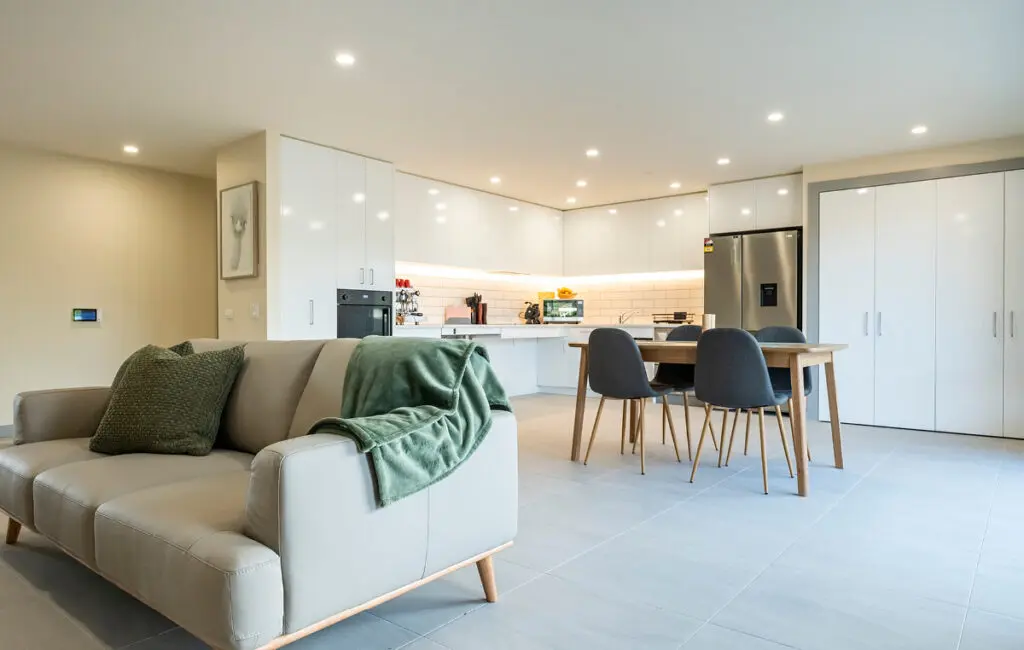 Open-plan living area with beige sofa, dining table and chairs, and white kitchen with stainless fridge.